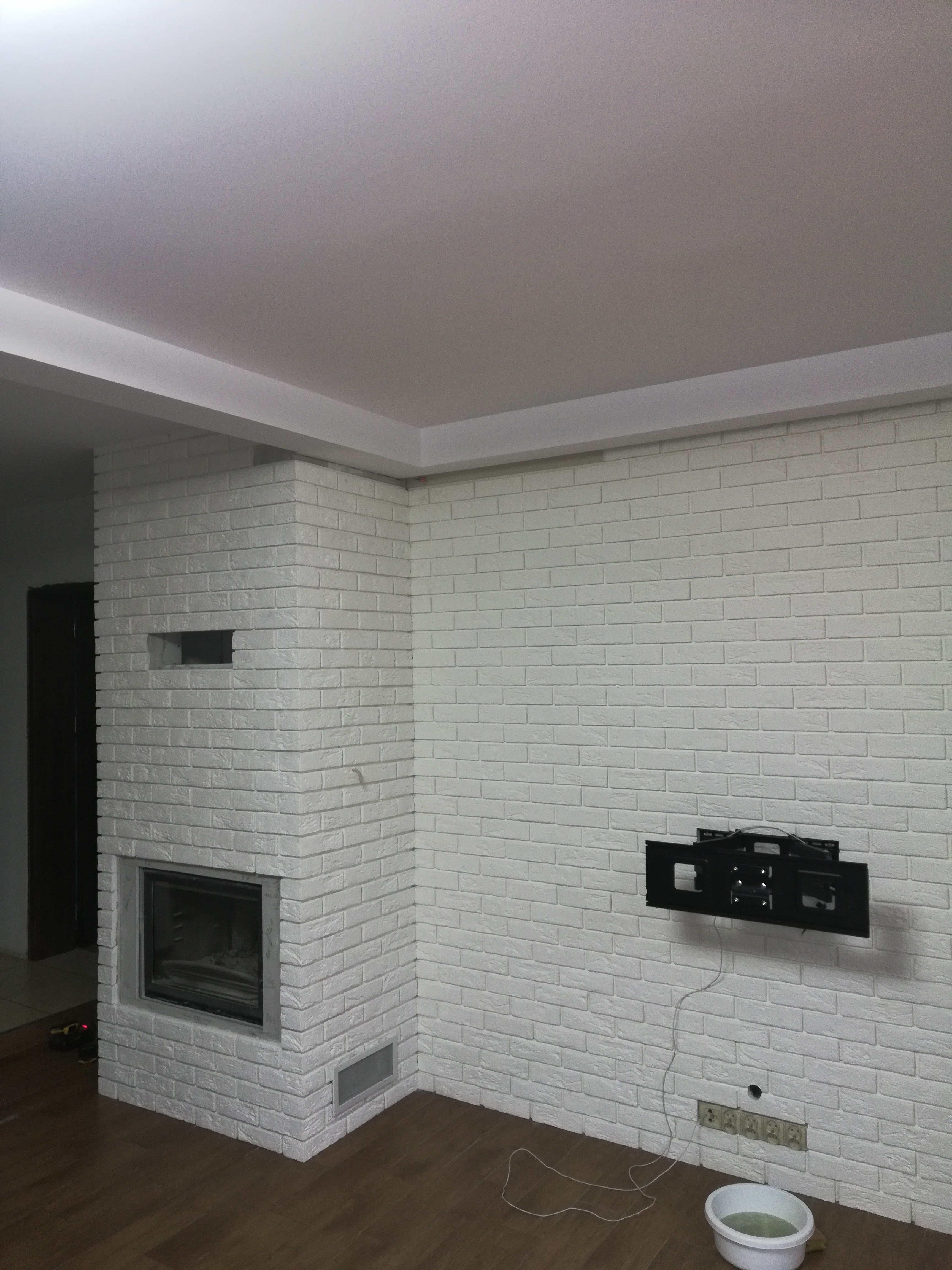 Ściana z cegły pomalowana na biało z zamontowanym uchwytem do telewizora, widoczne gniazdka elektryczne i miska z wodą na podłodze.