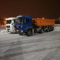 Dwa samochody ciężarowe MAN: biały i niebieski z pomarańczową naczepą, stojące na ośnieżonym parkingu nocą, oświetlone lampami ulicznymi.