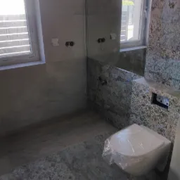 Nowoczesna łazienka w szaro-beżowych odcieniach z płytkami imitującymi kamień, wisząca toaleta zabezpieczona folią, widoczne puszki elektryczne na ścianie.