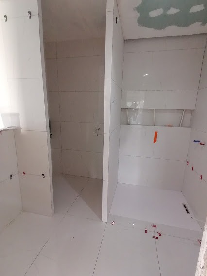 Białe płytki ceramiczne układane w dwóch kabinach prysznicowych, widoczne klipsy poziomujące i fragment surowego sufitu.