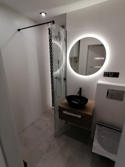 Nowoczesna łazienka z czarną umywalką nablatową, podświetlanym okrągłym lustrem i kabiną prysznicową w trakcie montażu, zabezpieczoną folią ochronną.