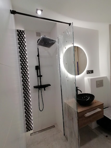 Nowoczesna łazienka z czarną armaturą, kabiną prysznicową typu walk-in z folią ochronną, okrągłym lustrem z podświetleniem LED i umywalką nablatową w kolorze czarnym.