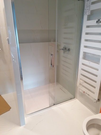 Nowoczesna kabina prysznicowa z przesuwnymi drzwiami, chromowanymi okuciami i białym grzejnikiem drabinkowym, widok na fragment toalety.