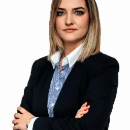 Profesjonalny portret kobiety w ciemnej marynarce i wzorzystej koszuli z założonymi rękami, patrzącej wprost, z delikatnym uśmiechem i złotym zegarkiem na nadgarstku.