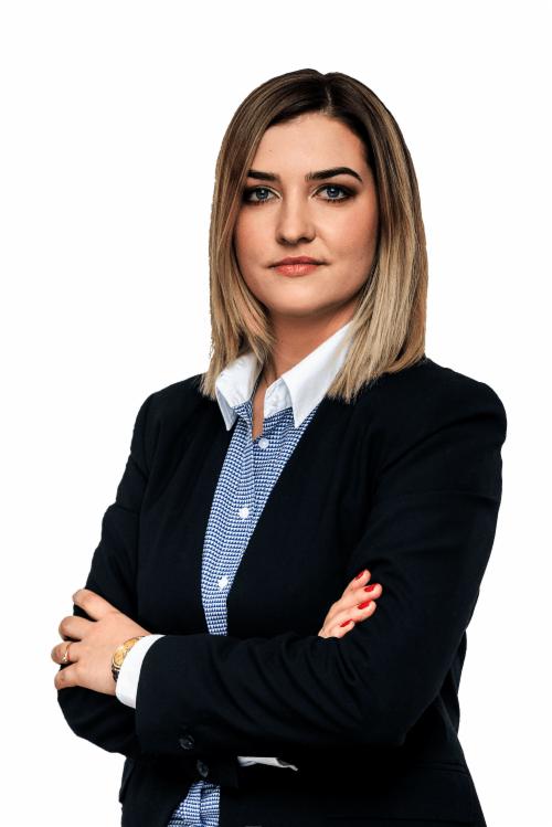 Profesjonalny portret kobiety w ciemnej marynarce i wzorzystej koszuli z założonymi rękami, patrzącej wprost, z delikatnym uśmiechem i złotym zegarkiem na nadgarstku.