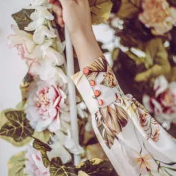 Kwiatowy mankiet bluzki z ozdobnymi guzikami, trzymany przez dłoń na tle kwiatowej dekoracji. Detal szycia z pracowni w Poznaniu.