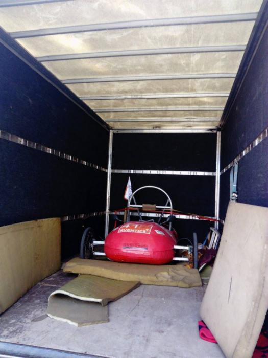 Czerwony gokart z naklejką 'Aventics' zabezpieczony piankowymi podkładkami wewnątrz ciemnej furgonetki transportowej z metalową ramą.