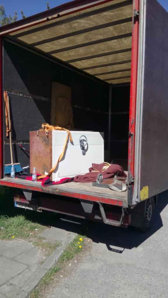 Wnętrze otwartej ciężarówki z widoczną skrzynią zabezpieczoną pasem, obok butelka wody i zmiotka, sugerujące przerwę w załadunku podczas transportu.