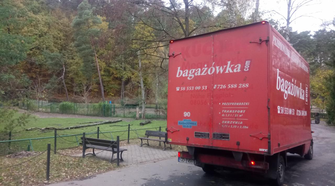 Czerwona furgonetka firmy przeprowadzkowej z logo 'bagażówka.com' zaparkowana na obrzeżach parku w jesiennej scenerii, widoczne ławki parkowe i drzewa w tle.