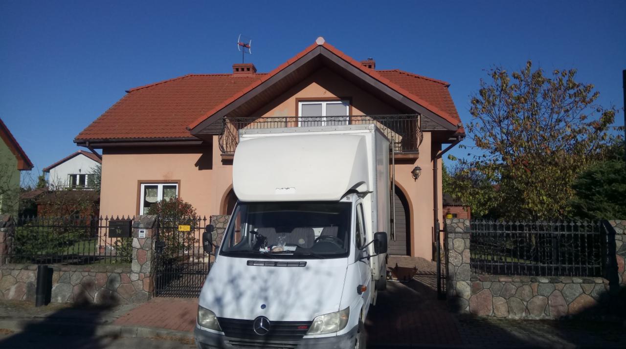 Biały samochód dostawczy Mercedes z wysoką zabudową zaparkowany przed domem z czerwonym dachem i balkonem, przygotowany do transportu rzeczy.