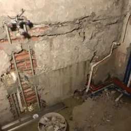 Surowy stan ściany z odsłoniętą cegłą i poprowadzonymi rurami instalacji wodnej z niebieskimi i czerwonymi oznaczeniami, widoczne ślady po kuciu i resztki gruzu na podłodze.