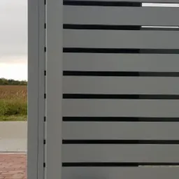 Szara brama przesuwna z poziomymi panelami, napędem elektrycznym i ż&oacute;łtą lampą ostrzegawczą na słupku, widok z boku na tle pola i nieba.