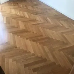 Ułożona w jodełkę, jasna, drewniana podłoga w pustym, jasnym pomieszczeniu.