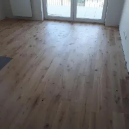 Jasna, szczotkowana podłoga z desek w nowym mieszkaniu, widoczne drzwi balkonowe z metalową barierką i grzejnik na ścianie.