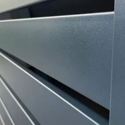 Szczegółowe ujęcie fragmentu nowoczesnej, szarej bramy wjazdowej z poziomych, metalowych profili, ukazujące fakturę i precyzję wykonania.