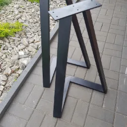 Dwie czarne, metalowe nogi do stołu z płaskownikami na górze z otworami na śruby, ustawione na szarej kostce brukowej, z ozdobnym kamiennym tłem i roślinnością.