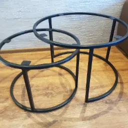 Dwie czarne, metalowe ramy stolików kawowych z okrągłymi blatami, wykonane ze spawanych rur, stojące na drewnianej podłodze.