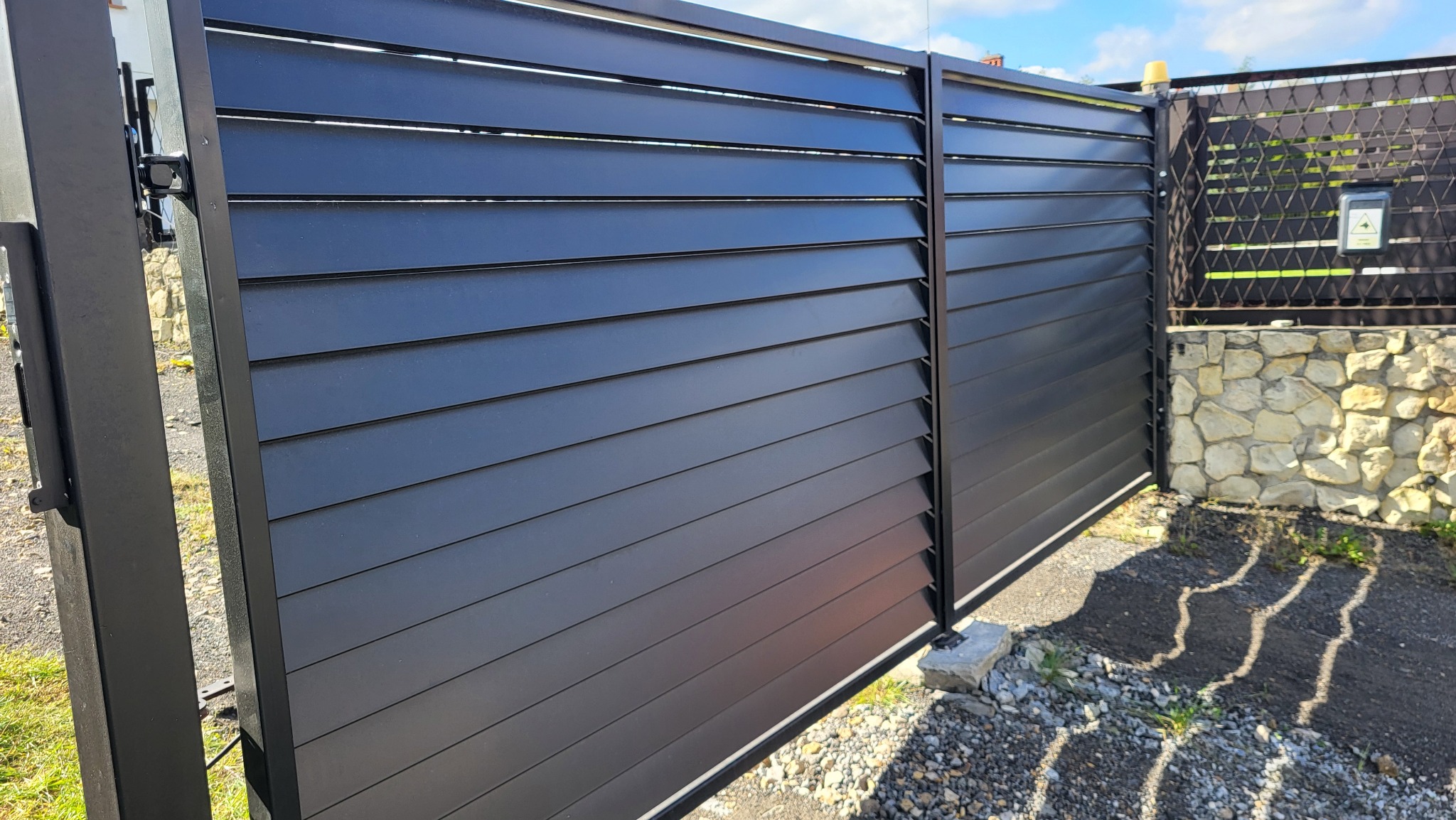 Nowoczesna, przesuwna brama wjazdowa z poziomych, czarnych paneli, widoczny zamek i fragment ogrodzenia z siatki na kamiennej podmurówce.