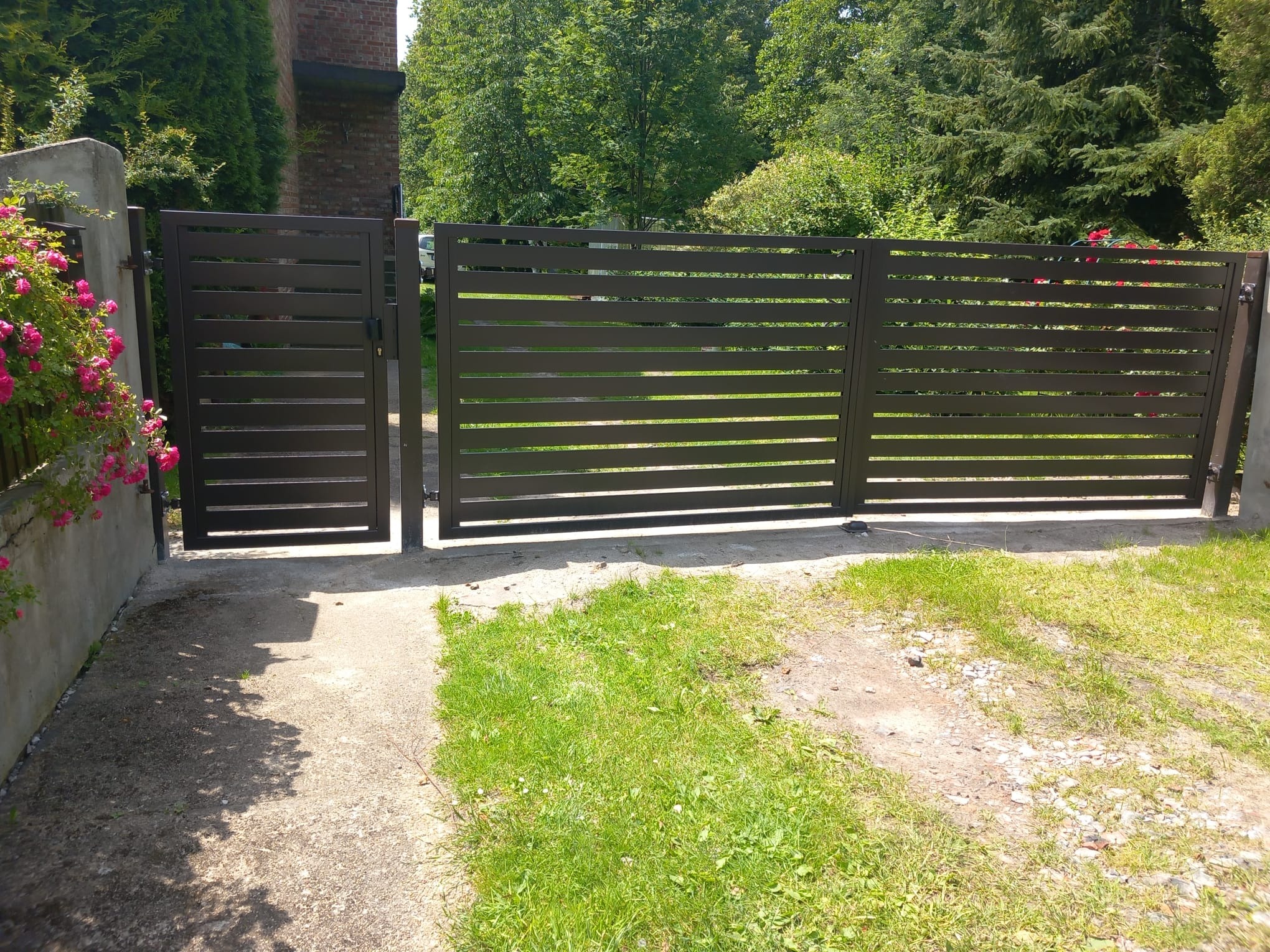 Nowoczesna, pozioma brama wjazdowa z metalowymi panelami w kolorze ciemnego brązu, z widoczną furtką po lewej stronie, na tle zieleni ogrodu i ceglanego budynku.