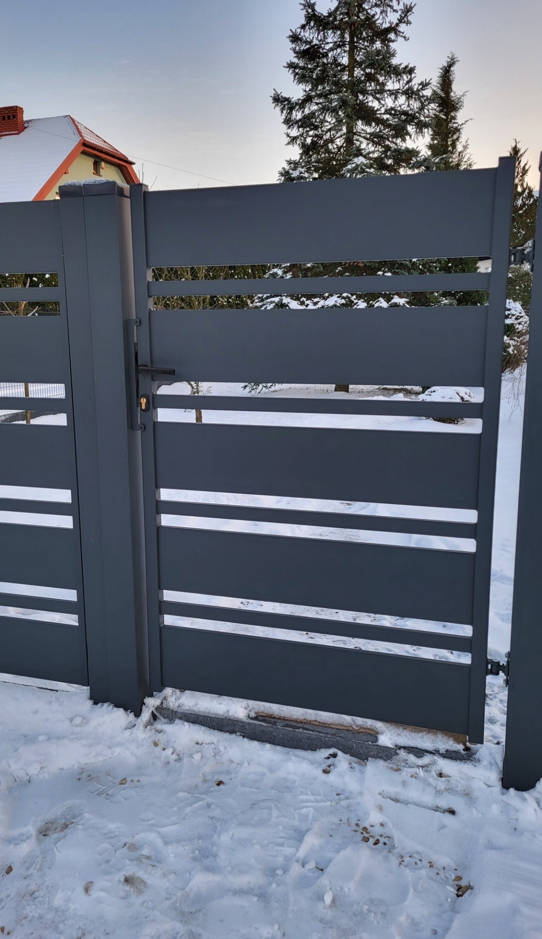 Nowoczesna, grafitowa brama wjazdowa z poziomymi prześwitami na tle zimowego krajobrazu z ośnieżonym domem w oddali.