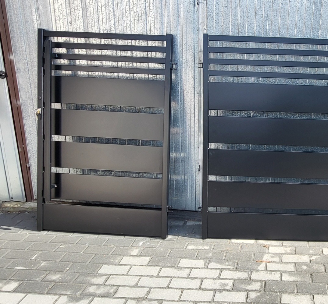 Dwie czarne, nowoczesne furtki z poziomymi prętami i dekoracyjnymi wstawkami z mozaiki, ustawione na kostce brukowej przed blaszakiem.