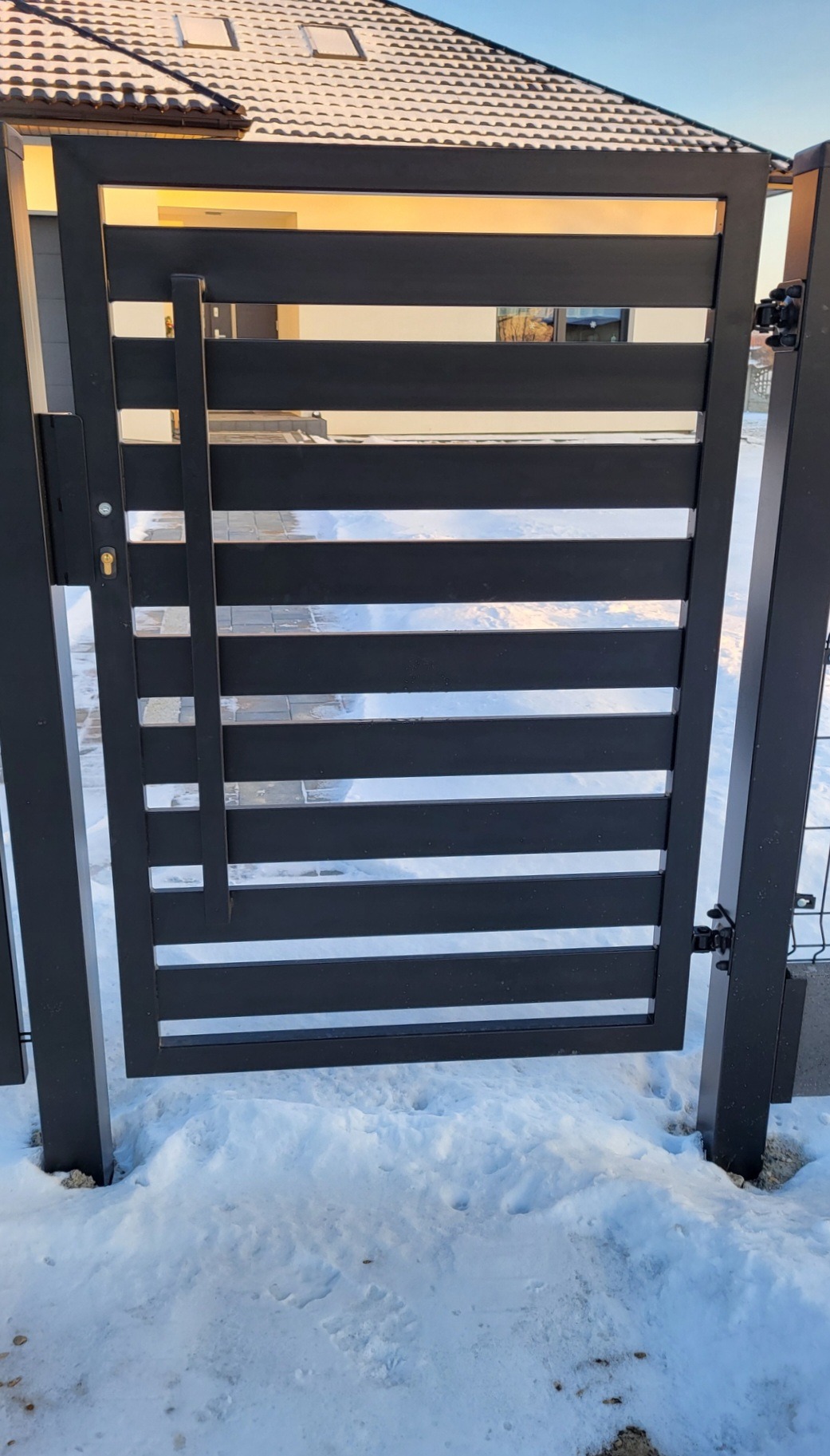 Nowoczesna, czarna brama wjazdowa z poziomych profili stalowych, zamontowana w Mstowie, widoczna na tle ośnieżonego podjazdu i jasnego budynku.