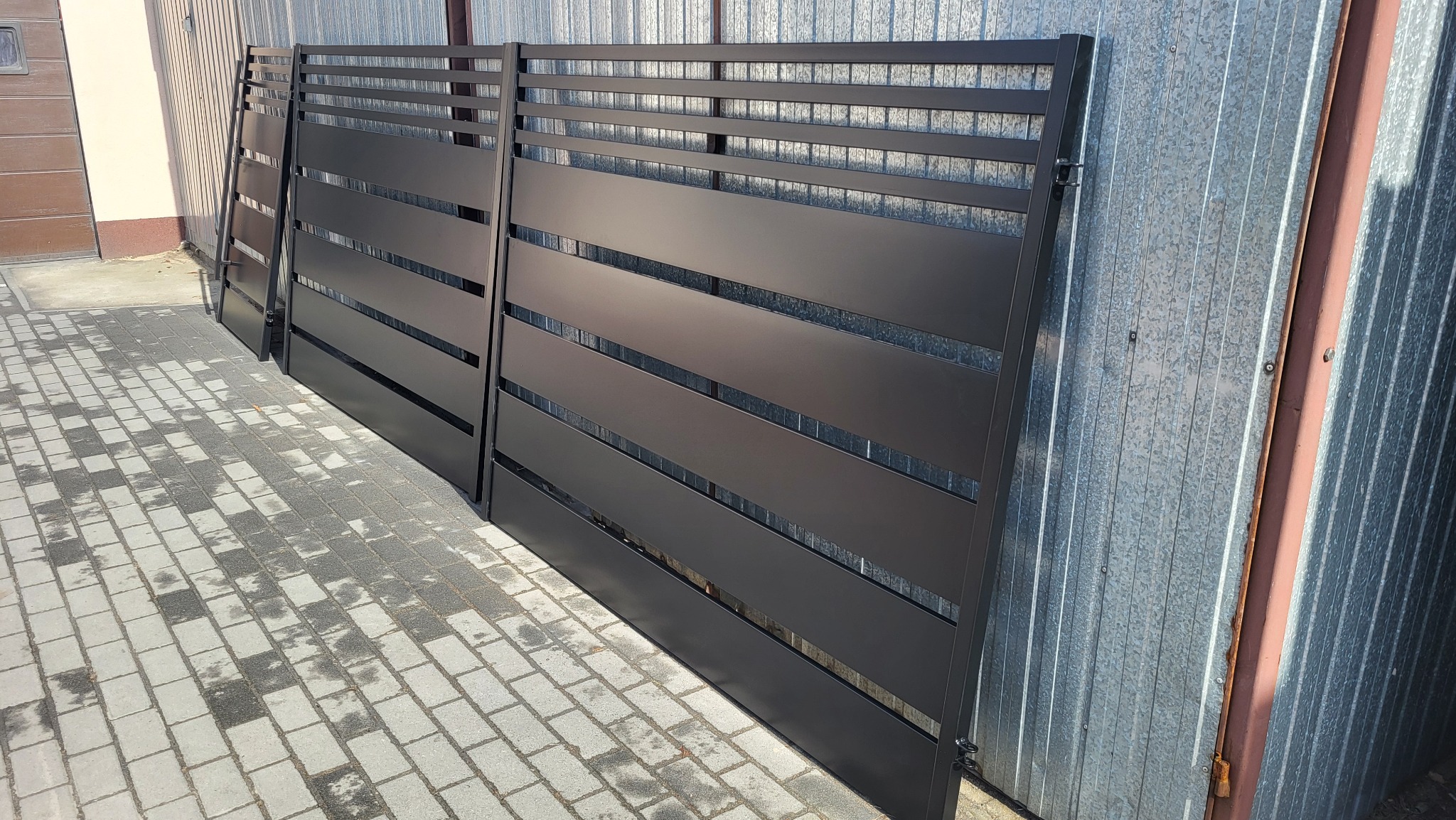 Nowoczesna, matowa, grafitowa brama wjazdowa z poziomymi panelami, oparta o blaszane ogrodzenie tymczasowe, na tle kostki brukowej.