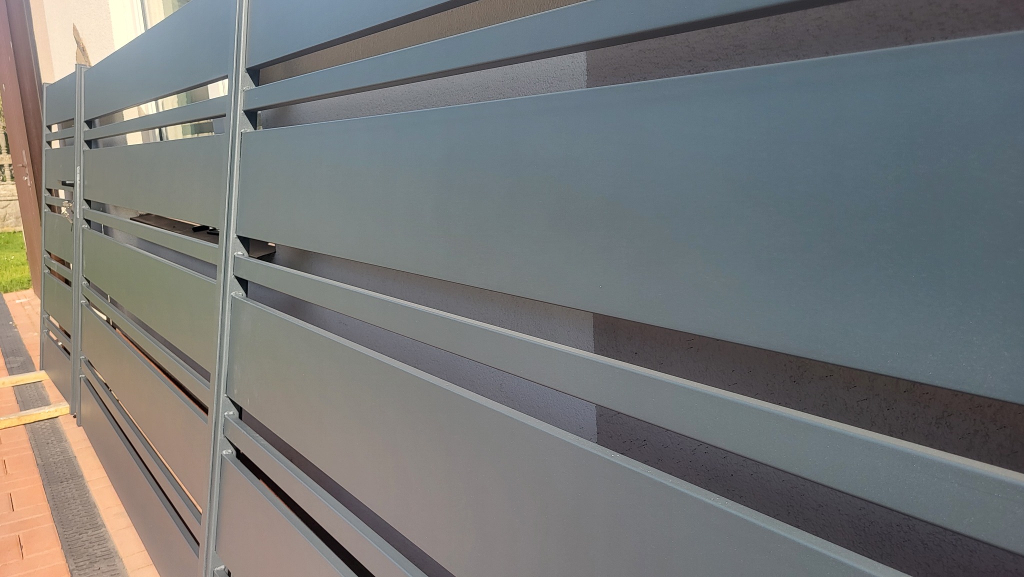 Nowoczesne, poziome ogrodzenie metalowe w kolorze szaro-zielonym, z widocznym fragmentem chodnika z kostki brukowej i trawnika w tle.