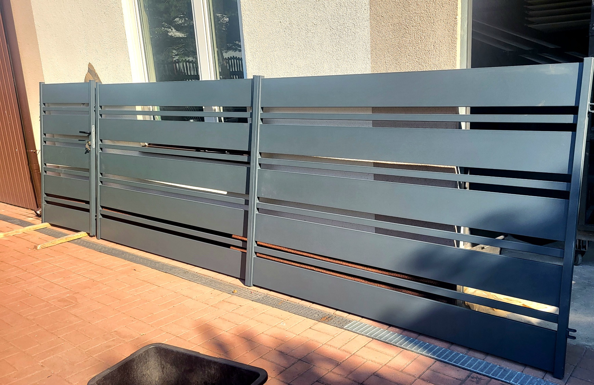 Nowoczesna, szara brama wjazdowa z poziomymi panelami, zamontowana na posesji z czerwoną kostką brukową, widoczne drewniane podpory i odpływ liniowy.