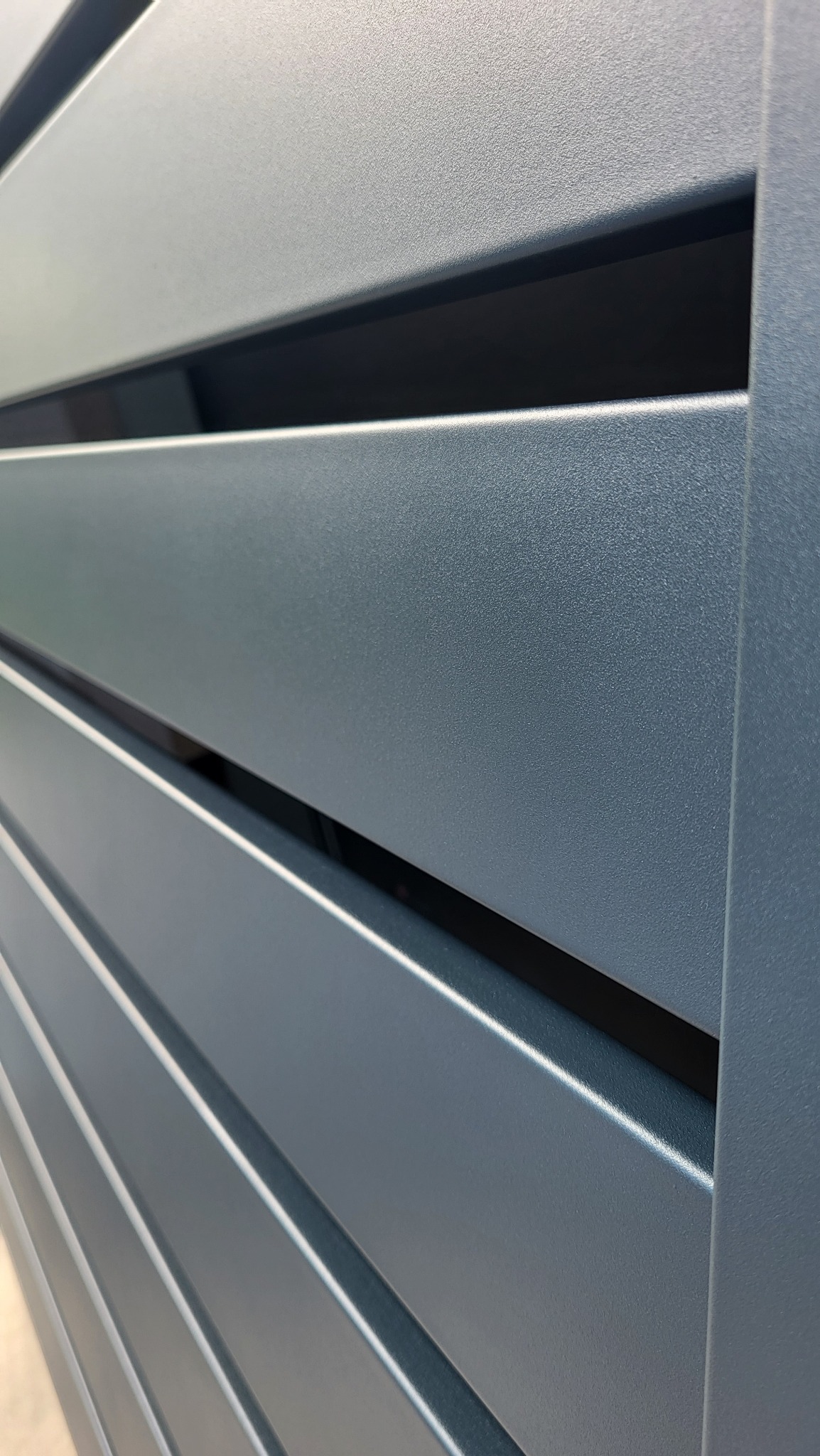 Szczegółowe ujęcie fragmentu nowoczesnej, szarej bramy wjazdowej z poziomych, metalowych profili, ukazujące fakturę i precyzję wykonania.