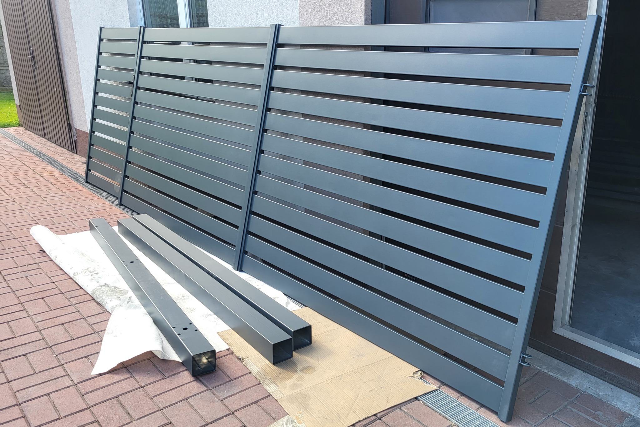 Elementy nowoczesnego, szarego ogrodzenia metalowego z poziomymi szczeblami, przygotowane do montażu na posesji wyłożonej czerwoną kostką brukową.