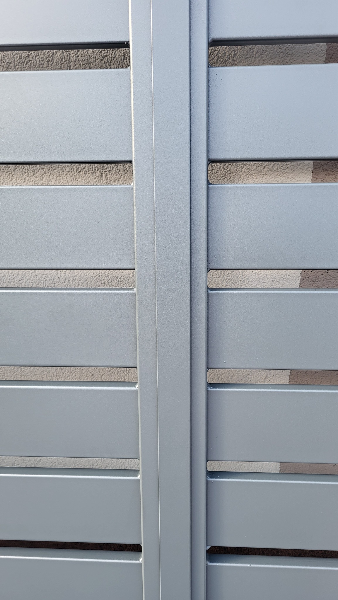 Fragment szarej, metalowej bramy wjazdowej z poziomymi szczeblami i widocznym fragmentem muru w tle