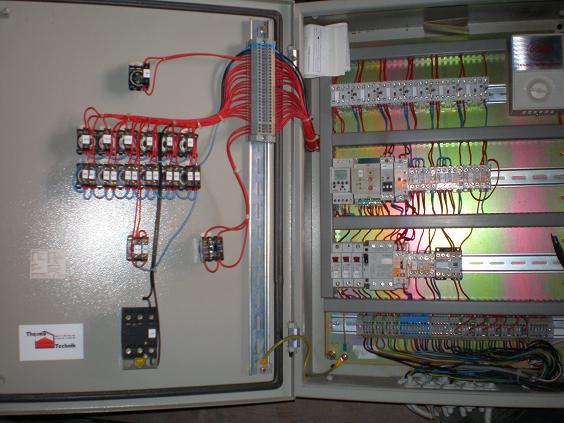 Otwarta szafa sterownicza z licznymi przekaźnikami, przewodami elektrycznymi w kolorach czerwonym, niebieskim i czarnym, oraz modułami sterującymi na szynach DIN, widoczny fragment logo firmy...