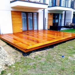 Qubix Sp.Z.o.o - Błyszczący, świeżo polakierowany taras drewniany przylega do białego domu z drewnianą elewacją. Na trawniku leżą narzędzia i ziemia, co sugeruje trwające prace wykończeniowe.