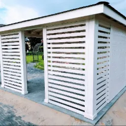 Biała, nowoczesna altana z poziomymi lamelami, widoczna z zewnątrz na tle domu i ogrodu. Kostka brukowa wokół konstrukcji.