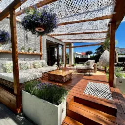 Drewniany taras z pergolą, osłonięty siatką cieniującą. Wygodne meble ogrodowe, donice z lawendą i wiszące kwiaty tworzą relaksującą przestrzeń.