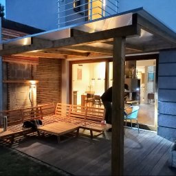 Qubix Sp.Z.o.o - Drewniana pergola z przezroczystym zadaszeniem przylegająca do domu, oświetlona lampą, z drewnianym zestawem wypoczynkowym na tarasie. Widok wieczorny.