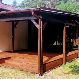 Qubix Sp.Z.o.o - Zadaszony taras drewniany z brązowymi elementami konstrukcyjnymi, przylegający do budynku. W tle widoczna altana z grillem i osoba siedząca.