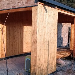 Qubix Sp.Z.o.o - Surowa drewniana konstrukcja z widocznymi elementami budynku w tle, ukazana w jasnym świetle słonecznym, z widocznymi śladami budowy.