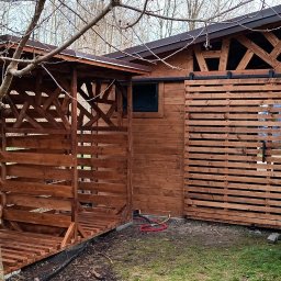 Qubix Sp.Z.o.o - Drewniana wiata na drewno z przesuwanymi drzwiami i dekoracją ścienną z motywem roślinnym. Konstrukcja z bejcowanego drewna na zewnątrz.