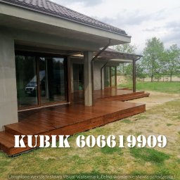 Qubix Sp.Z.o.o - Nowoczesny taras drewniany zadaszony pergolą, brązowe deski, widok z zewnątrz, szklane drzwi przesuwne, trawnik. Widoczny numer telefonu na desce.