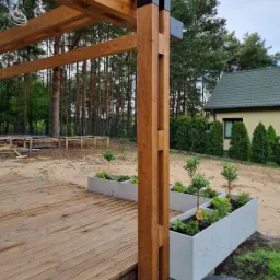 Drewniana pergola z czarnymi łącznikami, betonowe donice z roślinami, deski tarasowe. Widok na ogród z domem w tle. Ujęcie z dołu, podkreślające konstrukcję.