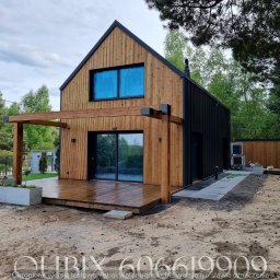 Qubix Sp.Z.o.o - Nowoczesny dom z drewnianą elewacją i tarasem, połączony z pergolą. Widoczne czarne panele, okna z niebieską folią, otoczenie leśne, świeżo zagospodarowany teren.