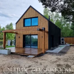 Nowoczesny dom z drewnianą elewacją i tarasem, połączony z pergolą. Widoczne czarne panele, okna z niebieską folią, otoczenie leśne, świeżo zagospodarowany teren.