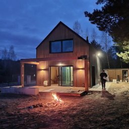 Qubix Sp.Z.o.o - Nowoczesny dom z drewnianą elewacją i pergolą oświetloną lampami, ognisko na pierwszym planie, osoba w tle. Ujęcie wieczorne.
