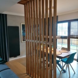 Qubix Sp.Z.o.o - Drewniana ścianka działowa z pionowych listew oddziela salon od jadalni. Widoczny stół z krzesłami i okno z roletami w tle. Ujęcie z niskiego kąta.