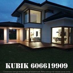 Qubix Sp.Z.o.o - Nowoczesny dom z drewnianym tarasem oświetlonym wieczornym światłem, widok z ogrodu. Duże okna z drewnianymi ramami. Widoczny fragment trawnika.