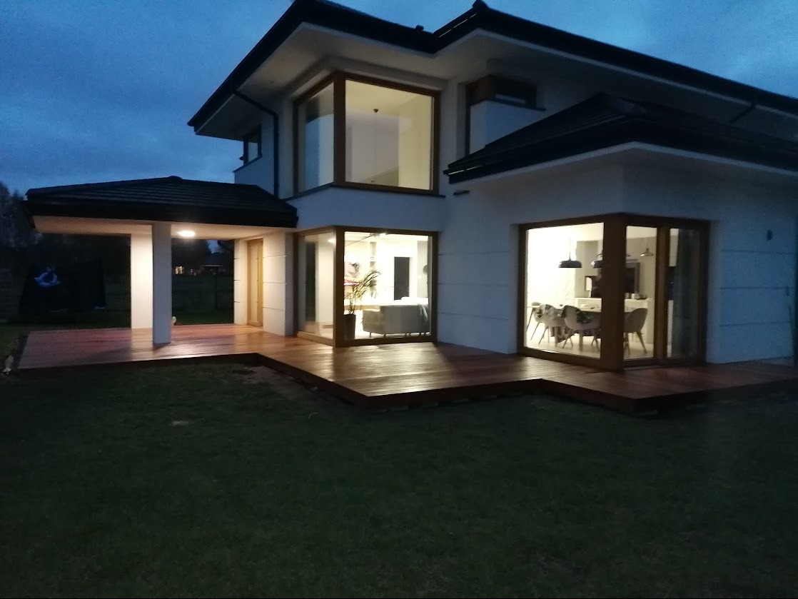 Nowoczesny dom z drewnianym tarasem i dużymi oknami, oświetlony wieczornym światłem. Widoczny fragment trawnika i zadaszony taras z kolumnami.