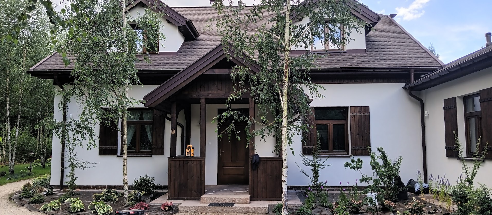 Biały dom z brązowym dachem i drewnianymi elementami, otoczony zielenią. Widoczne drewniane okiennice, wejście z zadaszeniem i fragment ogrodu.