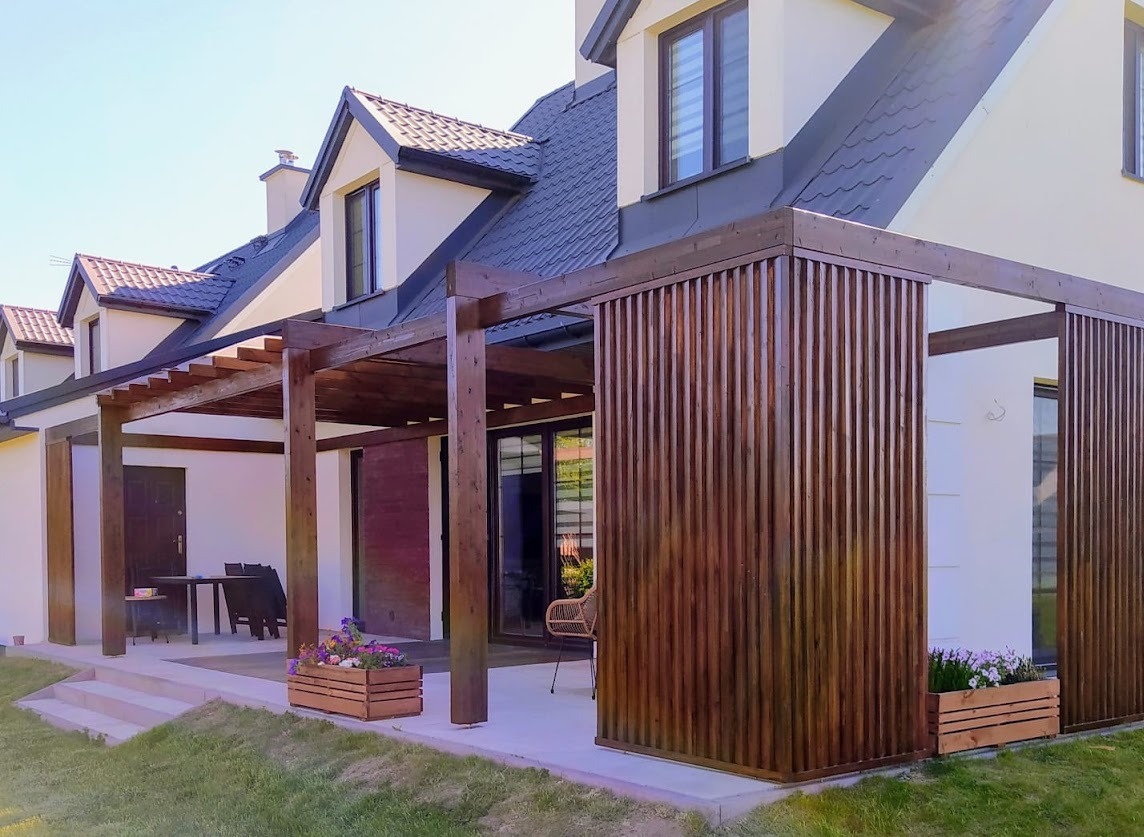 Drewniana pergola z pionowymi panelami osłaniającymi część tarasu, z donicami z kwiatami. Dom z ciemnym dachem w tle.